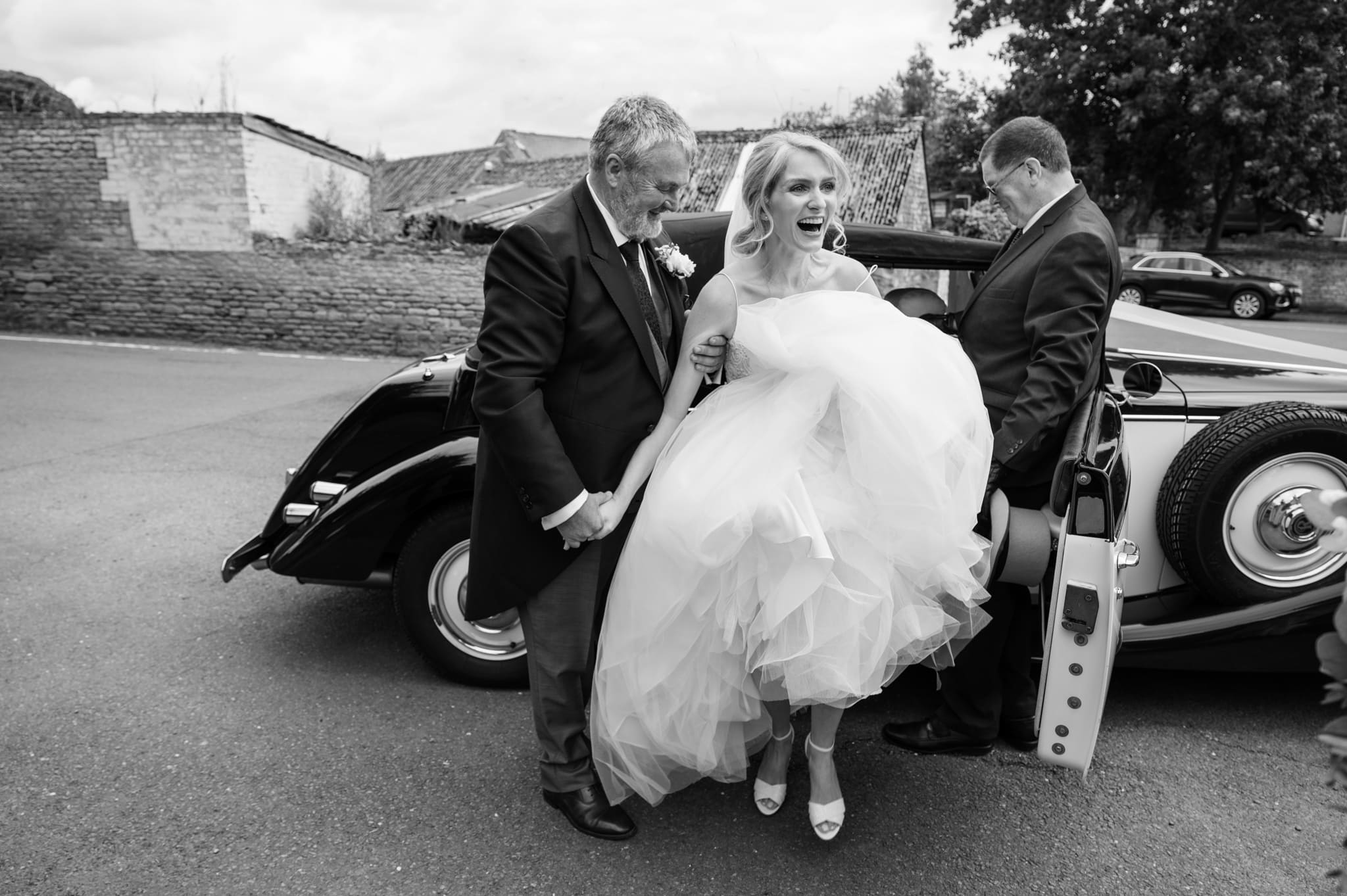 Bride laughing as she tries to get out of the wedding car with her huge dress