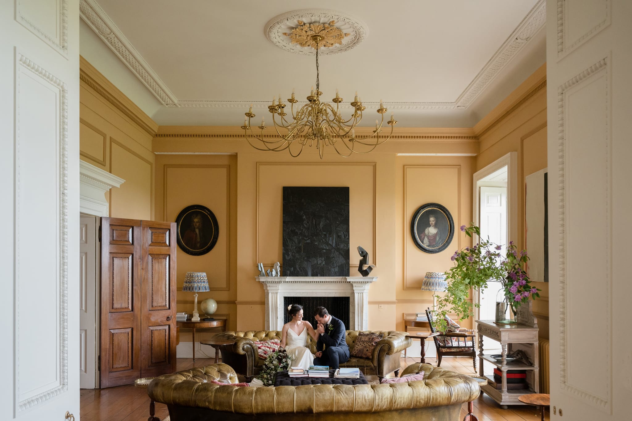 Groom kissing back of bride's hand as they sit on a round chesterfield sofa