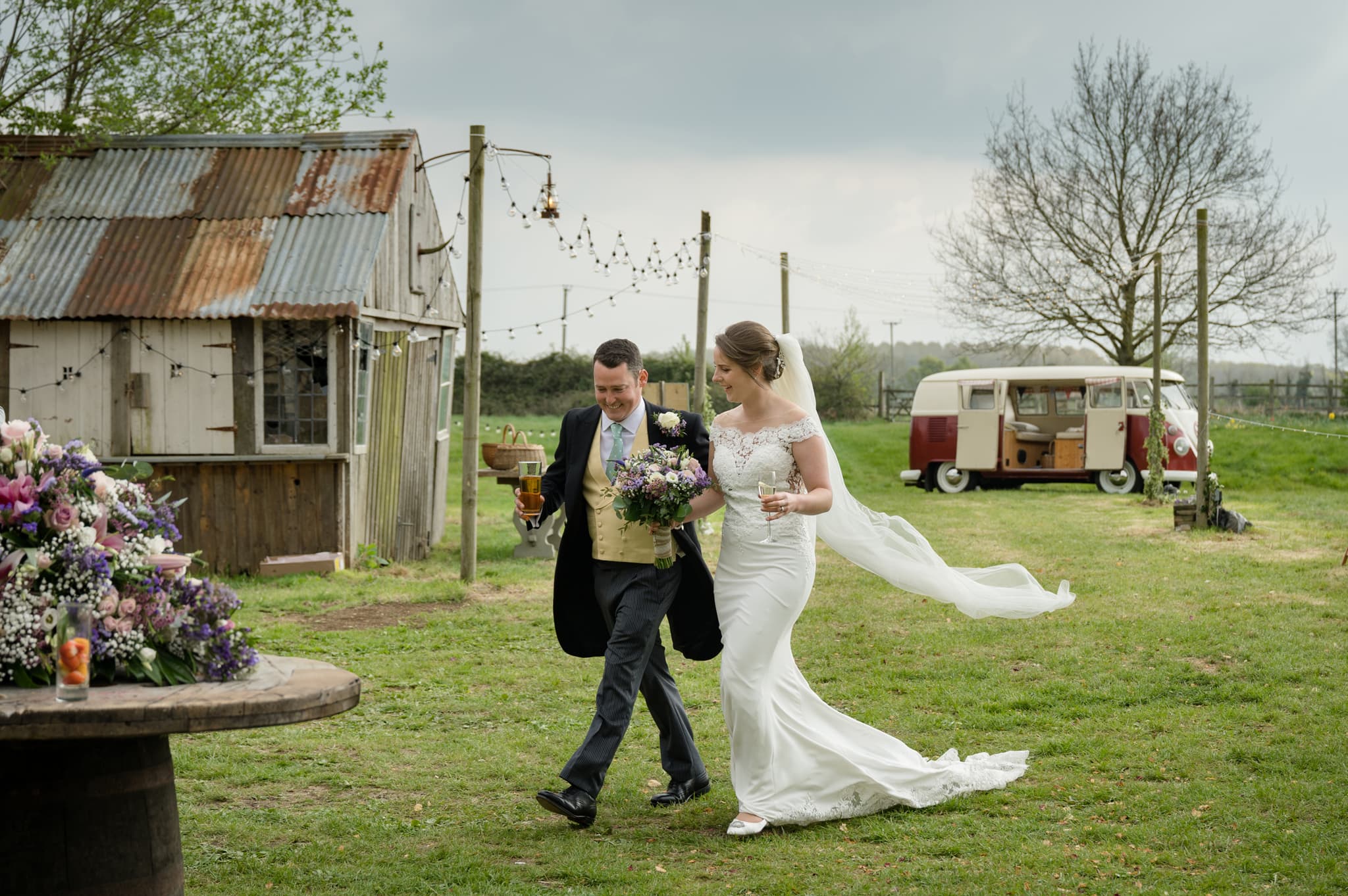 Bride and groom walking through a village hall field with their campervan in the background