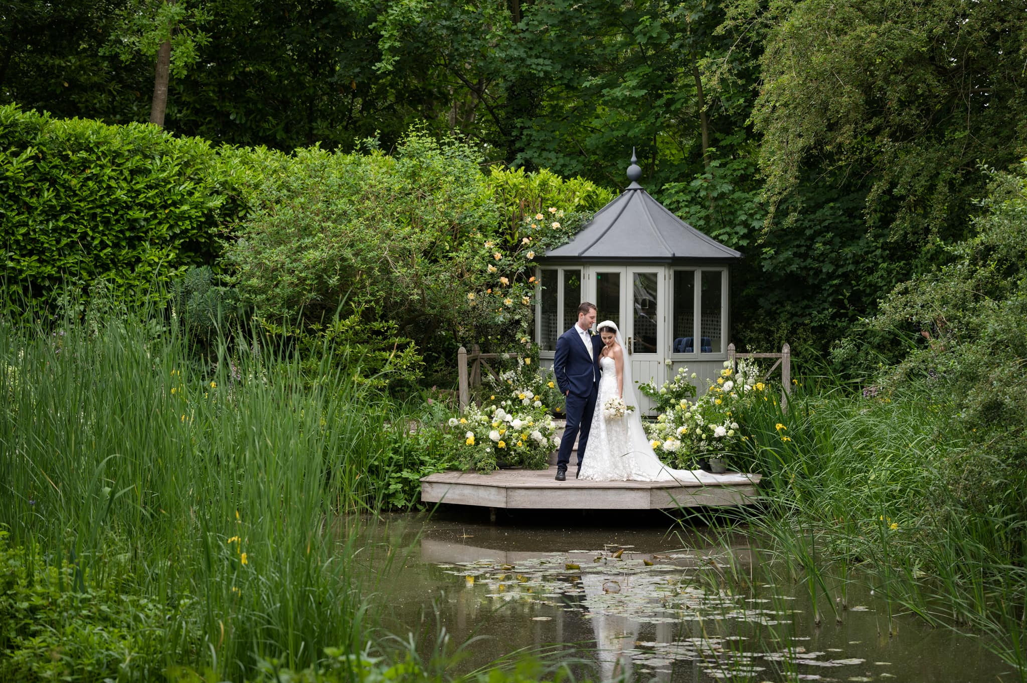 The bride and groom cuddled up on a wooden jetty over a pond with a summerhouse behind them