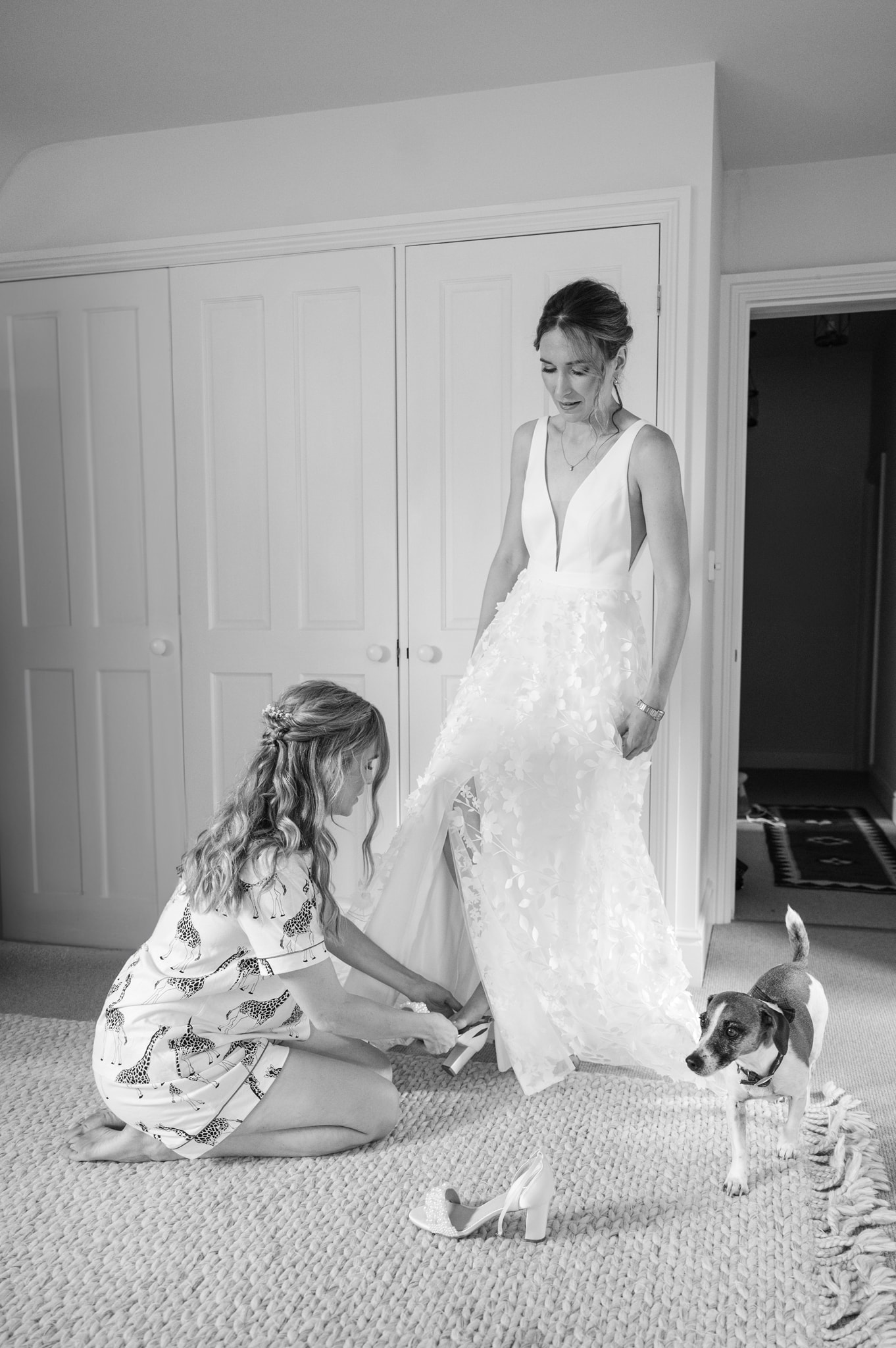 A bridesmaid putting the bride's shoes on for her as a little Jack Russell dog walks by