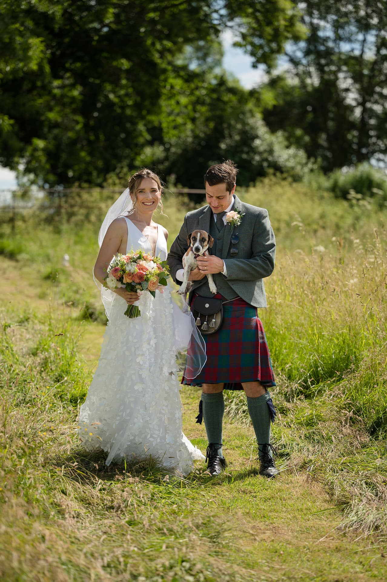 Groom holding a Jack Russell dog as he walks through a field with the bride