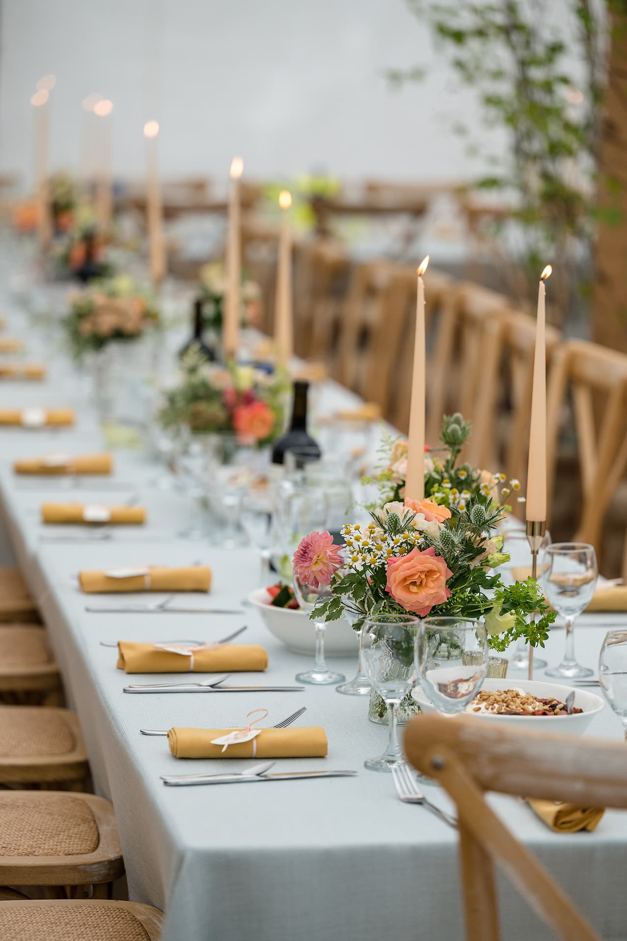 A dinner table set with pale blue linen, mustard napkins, vases of flowers, and cream candles.