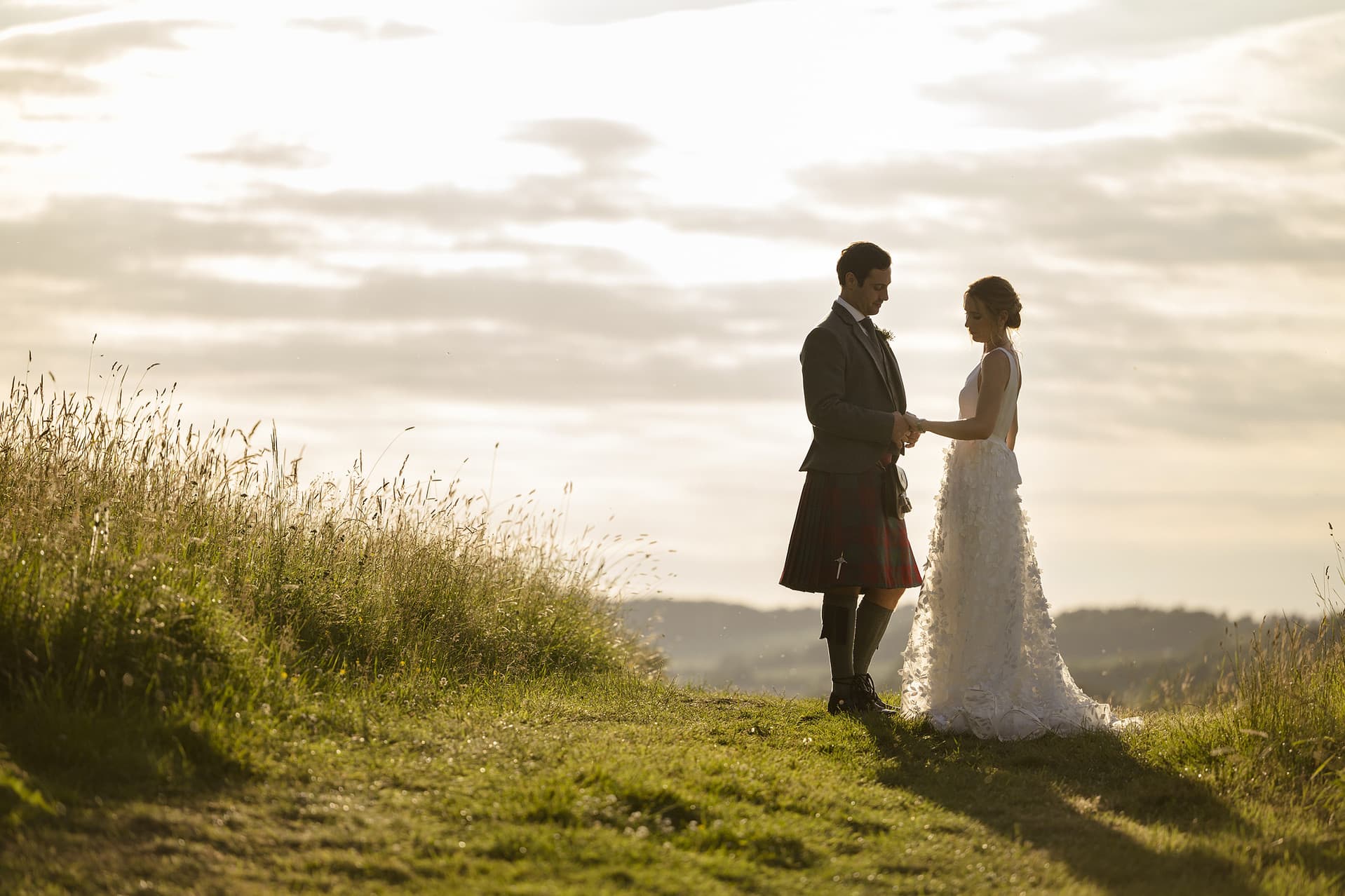 Groom looking at the bride's wedding ring as they stand in a field during golden hour