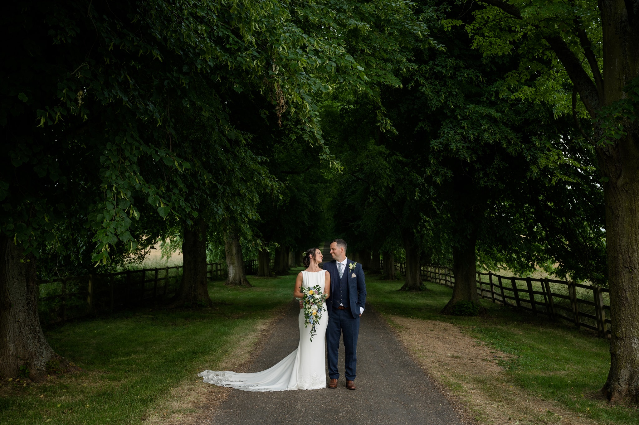 The bride and groom posing for a photo on the tree-lined avenue at Notley Abbey