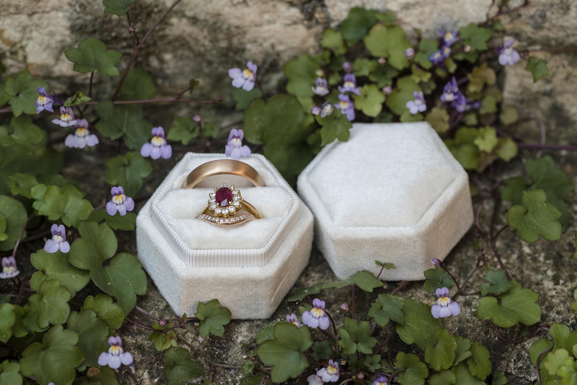 Wedding rings in a cream velvet octagonal box surrounded by tiny purple viola flowers on a stone wall at Notley Abbey